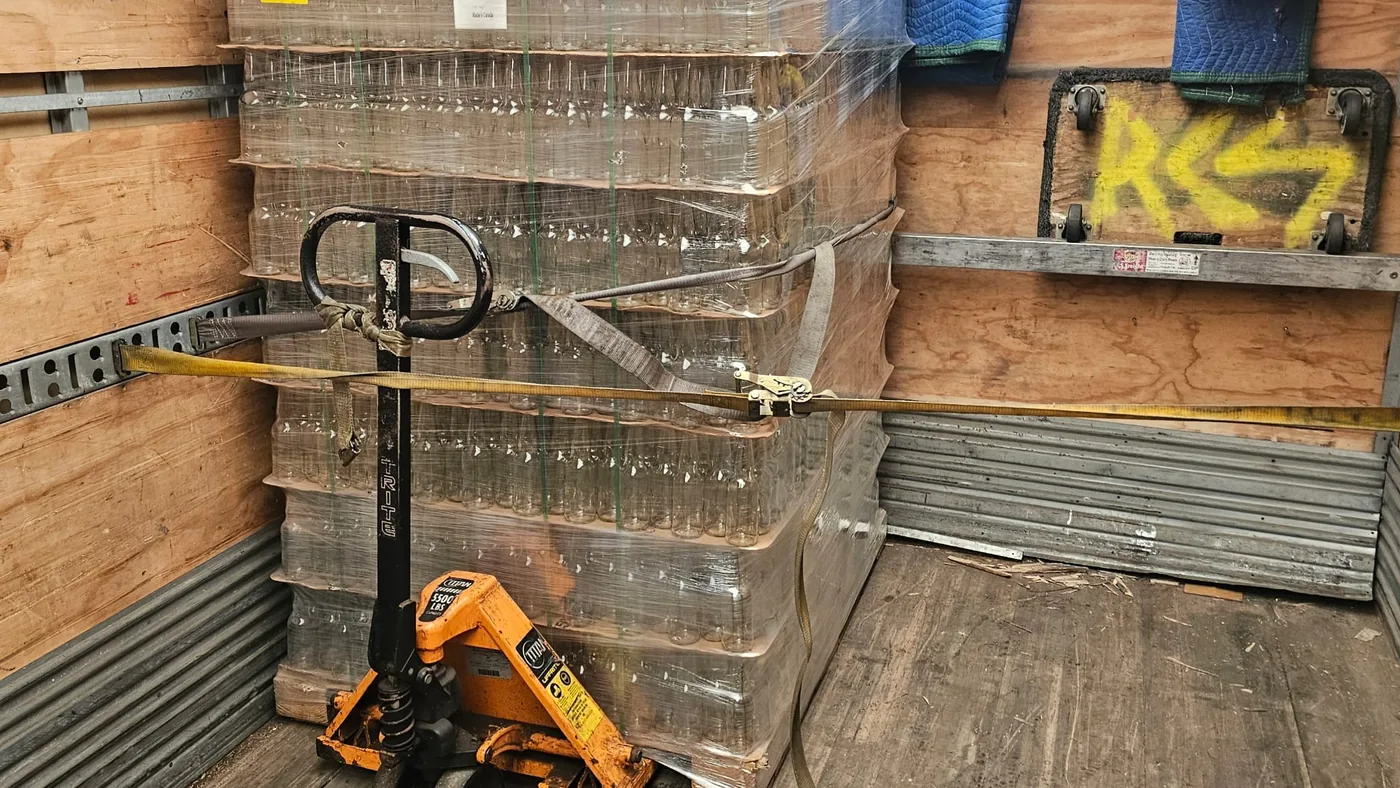 A shrink-wrapped pallet of glass bottles strapped to the wall of a Bravo 24 truck, pallet jack alongside.